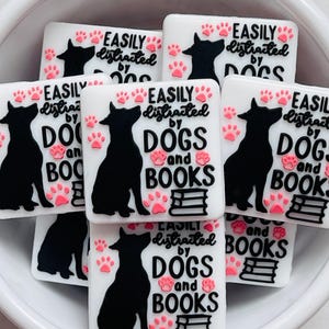 May include: White rectangular charms with a black dog silhouette, pink paw prints, and the text "EASILY distracted by DOGS and BOOKS." The charms are in a white bowl.