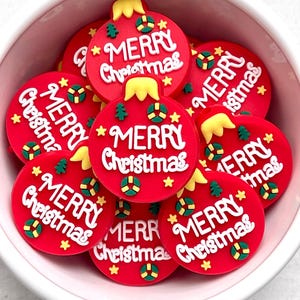May include: A collection of red Christmas ornament-shaped decorations with the words "MERRY Christmas" in white. Each ornament has yellow, green, and gold accents. The ornaments are inside a white bowl.