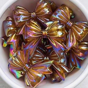 May include: A collection of iridescent, bow-shaped embellishments in a white bowl. The bows have a metallic, golden-brown hue with hints of rainbow iridescence. These decorative bows are likely used for crafts or embellishments.