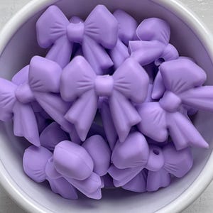 May include: A close-up shot of a white bowl filled with numerous lavender-colored bow-shaped beads. The beads are clustered together, showcasing their detailed bow design. The bowl is set against a white background.