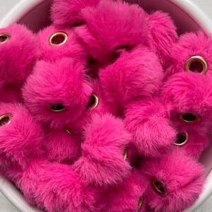 May include: A collection of vibrant fuchsia pink pom-poms with gold eyelets, filling a white bowl. The fluffy, textured pom-poms are densely packed, creating a visually appealing display. These craft supplies are ideal for various DIY projects.
