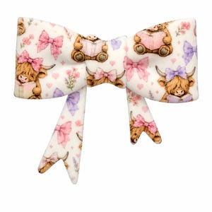 May include: A white fabric hair bow featuring a repeating pattern of brown highland cows and teddy bears reading books, accented with pink and purple bows, small hearts, and floral sprigs. The bow has two long, dangling tails with the same print.