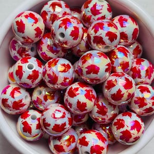 May include: A collection of round beads with a white base and a pattern of red maple leaves. The beads are contained in a white bowl. The beads have a hole through the center for stringing.