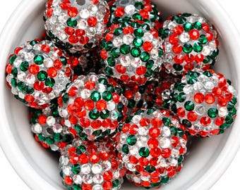 May include: A collection of spherical beads covered in small, faceted red, green, and clear crystals. Each bead features a central hole and is contained within a plain white bowl, showcasing a dense, sparkling texture across the rounded surfaces.