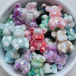 May include: A collection of iridescent bear-shaped beads in a white bowl. The beads are in various colors, including pink, purple, blue, and green. The beads have a glossy finish and are small in size.