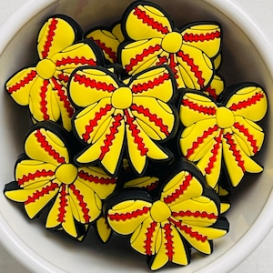 May include: A collection of yellow bow-shaped charms with red and black accents, nestled in a white bowl. The charms have a detailed design with a central knot and layered bow loops. The bright colors and playful shape suggest a craft or accessory item.