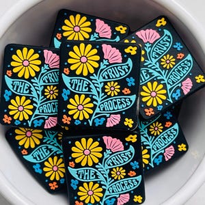May include: Black square magnets with a floral design and the words "Trust the Process." The design features yellow and pink flowers with blue and orange accents. The magnets are piled in a white bowl.