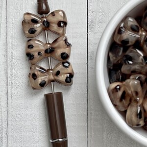 May include: A brown pen with three bow-shaped charms. The charms are a light brown color with black spots. The pen has a silver band near the bottom and a dark brown cap.