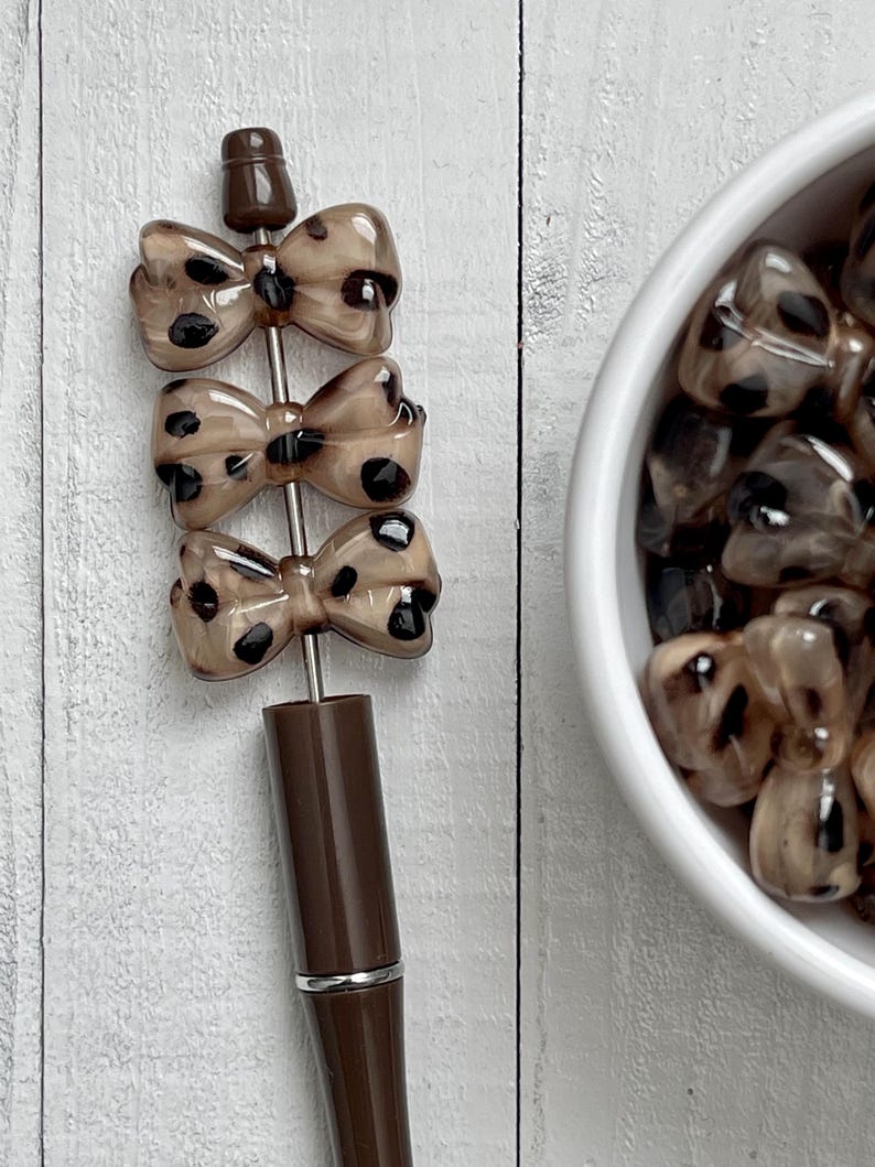 May include: A brown pen with three bow-shaped charms. The charms are a light brown color with black spots. The pen has a silver band near the bottom and a dark brown cap.