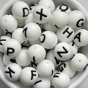 May include: A collection of white, round beads with black letters and symbols. The beads are inside a white bowl. The letters include D, X, 1, C, H, A, P, F, Z, and G. These beads are likely used for crafting or jewelry making.