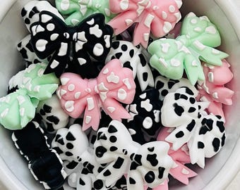 May include: A collection of decorative bow embellishments in a white bowl. The bows are in various colors, including black and white, pink and white, and mint green and white. The bows have a spotted pattern.