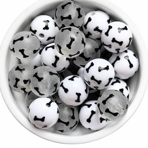 May include: A collection of round beads in a white bowl. The beads are a mix of opaque white and translucent gray, each featuring a repeating black bone pattern printed on the surface. The beads are spherical with a small hole visible in some.