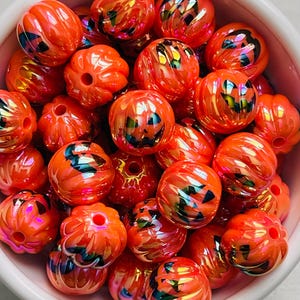 May include: A collection of orange, pumpkin-shaped beads with carved jack-o'-lantern faces. The beads have a glossy finish and iridescent highlights, creating a festive look. These beads are perfect for Halloween-themed crafts and jewelry making.