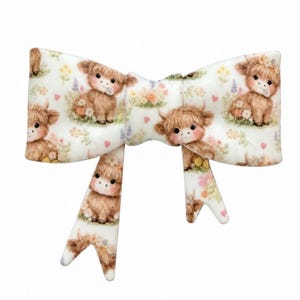 May include: A white fabric hair bow featuring a repeating pattern of brown Highland cattle calves sitting in patches of colorful wildflowers and small pink hearts. The bow has two long, pointed tails hanging down from the center knot.