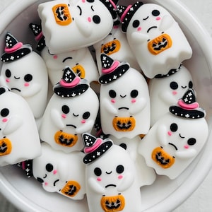 May include: A collection of white ghost-shaped decorations with black eyes, rosy cheeks, and small orange pumpkin-shaped bags. Each ghost wears a black witch hat with pink trim and white stars. The decorations are in a white bowl.