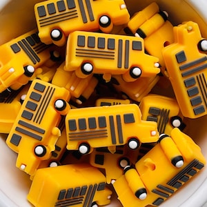 May include: A collection of yellow school bus charms with black and white details. The miniature buses have rectangular windows and are piled in a white bowl. The wheels are white with black centers.
