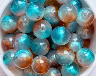 May include: A white bowl filled with iridescent beads. The beads are a mix of teal and copper colours, with a shimmering, translucent appearance. Each bead has a small hole, suggesting they are for stringing.