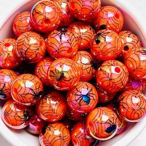 May include: A collection of orange, spherical beads with black spiderweb designs and small black spiders. The beads are glossy and appear to be made of glass or a similar material. They are displayed in a white bowl.