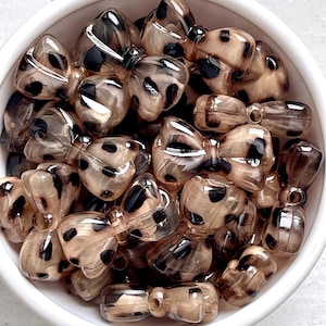 May include: A collection of bow-shaped beads in a white bowl. The beads are transparent with a tan base and black spots, resembling a leopard print. The beads are likely for jewelry making or crafting.