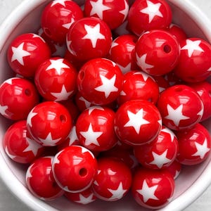 May include: A collection of glossy red beads with white star designs, each with a hole for stringing. The beads are round and uniform in size, filling a white bowl. Ideal for jewelry making or craft projects.
