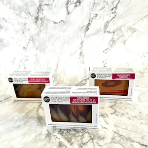 May include: Three bars of shea soap with different scents. The soaps are wrapped in white boxes with black and red text. The scents are Sun Seeker Peach Bergamot Cashmere, Cranberry Orange, and Orange Ginger Plum. The boxes are sitting on a white marble surface.