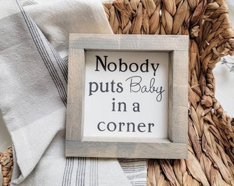 May include: A square wooden sign with a light gray stained frame and a white center panel. The text "Nobody puts Baby in a corner" is printed in black lettering on the white background. The sign is positioned within a woven basket.