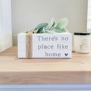 Puede incluir: Pila de libros decorativos blancos con el texto "There's no place like home" y un símbolo de corazón. Los libros están atados con hilo y cubiertos con hojas verdes y grises. A la derecha hay una vela perfumada con tapa negra.