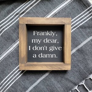 May include: A small, square wooden sign with the quote "Frankly, my dear, I don't give a damn" in white lettering on a black background. The sign is framed in light brown wood and rests on a gray and white striped fabric.