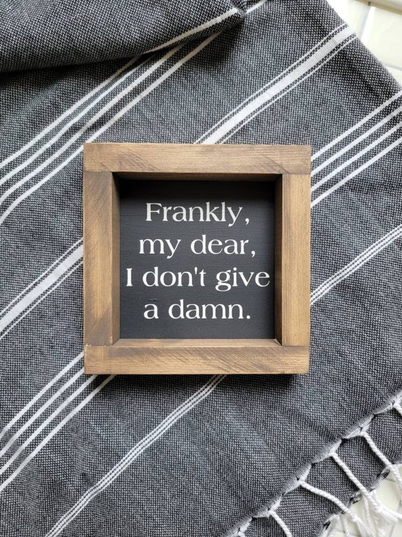 May include: A small, square wooden sign with the quote "Frankly, my dear, I don't give a damn" in white lettering on a black background. The sign is framed in light brown wood and rests on a gray and white striped fabric.