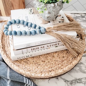 May include: A decorative tray holds books, a blue beaded garland, and a small concrete planter with greenery. The tray is woven from natural fibers. The books have white covers and titles like "Homebody" and "House Story".