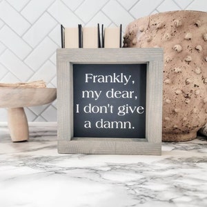 May include: A framed sign with the quote "Frankly, my dear, I don't give a damn." The sign has a gray wooden frame and a black background with white text. The sign is on a marble surface.