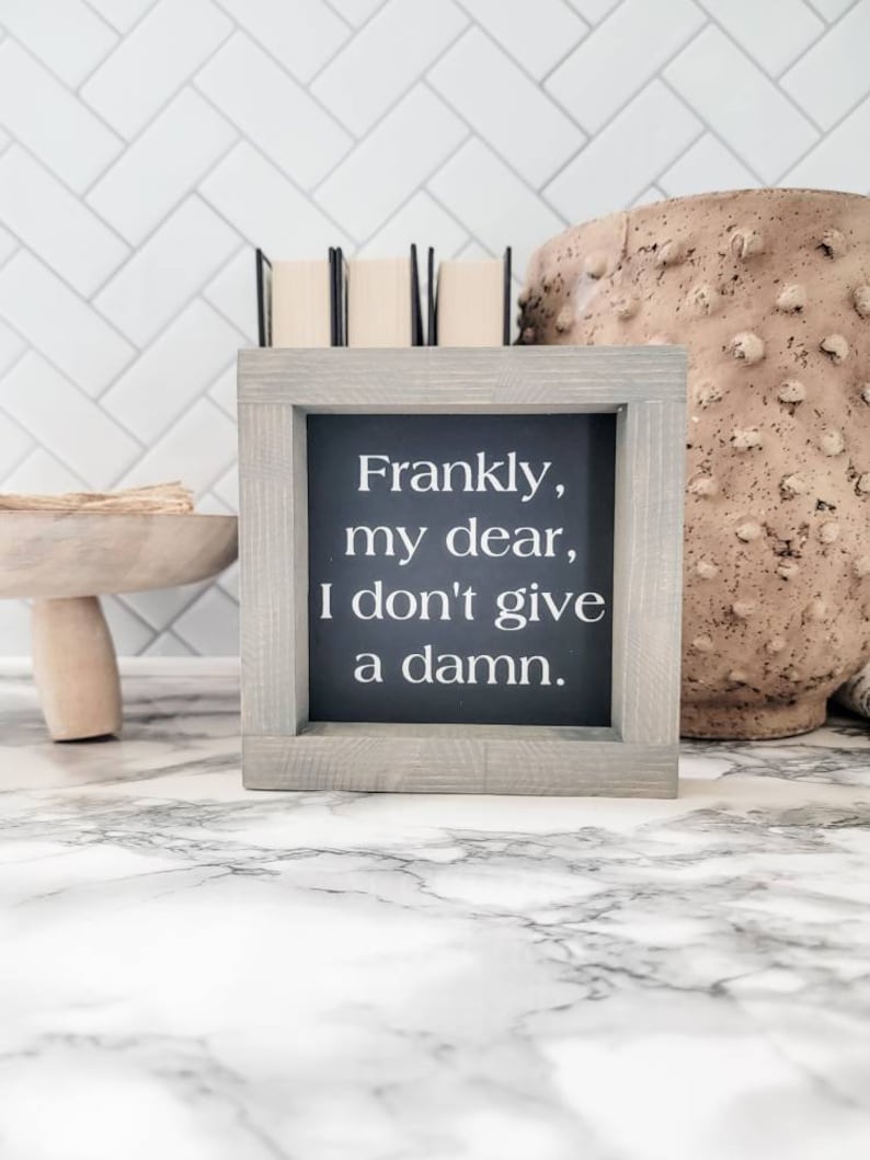 May include: A framed sign with the quote "Frankly, my dear, I don't give a damn." The sign has a gray wooden frame and a black background with white text. The sign is on a marble surface.