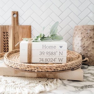May include: A stack of three white decorative books tied with twine. The top book reads "Home" with a heart symbol, followed by coordinates. A sprig of greenery rests on top. The books sit on a woven tray.