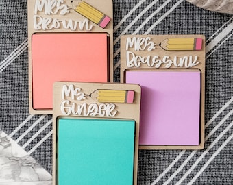 May include: Three wooden sticky note holders with colorful pads. Each holder has a name and a pencil design. The names are "Mrs. Brown", "Mrs. Doussin", and "Ms. Ginger". The pads are coral, lavender, and teal.