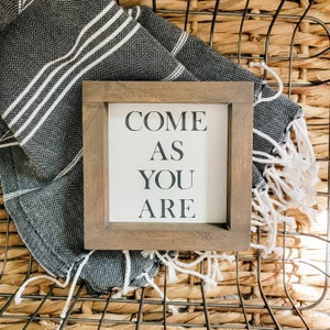 May include: A square wooden framed sign with the words "COME AS YOU ARE" in black lettering. The sign rests on a folded black and white striped towel with white fringe, set against a woven basket background.