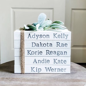 May include: A white wooden block decor piece with five stacked layers, each bearing a name in dark gray lettering: Adyson Kelly, Dakota Rae, Korie Reagan, Andie Kate, and Kip Werner. The piece is adorned with faux greenery and tied with twine.