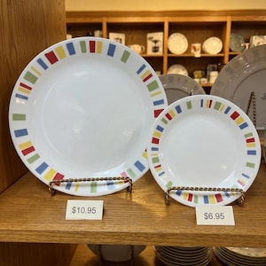 May include: Two white ceramic plates of different sizes, each featuring a decorative border of small, evenly spaced rectangular blocks in alternating colors of red, yellow, blue, and light green, displayed upright on gold-toned wire stands.
