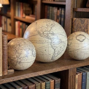 May include: Three antique-style globes of varying sizes, each with a cream-colored surface and detailed cartography. The globes are displayed on a wooden shelf alongside a collection of vintage books, creating a classic, scholarly aesthetic.