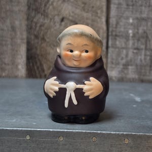 May include: A small ceramic figurine of a monk with a bald head, rosy cheeks, and a smiling expression. The figure wears a dark brown hooded robe with a white knotted rope belt around the waist, and has black shoes visible at the base.