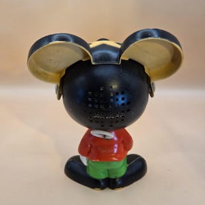 May include: Vintage Mickey Mouse figurine, viewed from the back. The figure has a black head with yellow ears, a red shirt, green shorts, and black shoes. The back of the head has a grid of small square holes.