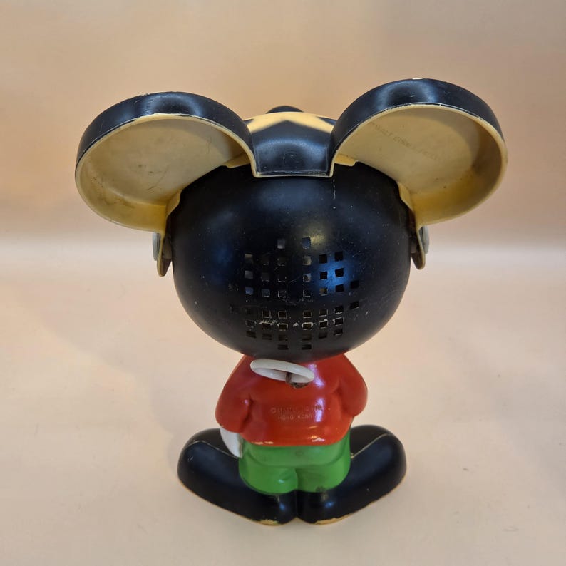 May include: Vintage Mickey Mouse figurine, viewed from the back. The figure has a black head with yellow ears, a red shirt, green shorts, and black shoes. The back of the head has a grid of small square holes.