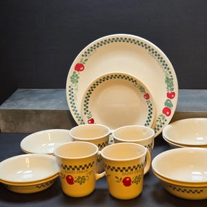 May include: A cream-colored dinnerware set featuring a green and black checkered border, red apple illustrations, and green grape clusters. The pieces include plates, bowls, and mugs, with the text "FARM FRESH" printed on the mugs and plates.