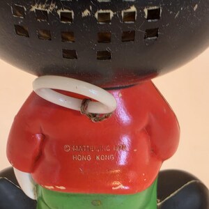 May include: Vintage toy figure with a black head and a red jacket. The figure has a white ring around its neck and green pants. The text "&copy; MATTEL, INC. 1976 HONG KONG" is printed on the red jacket.