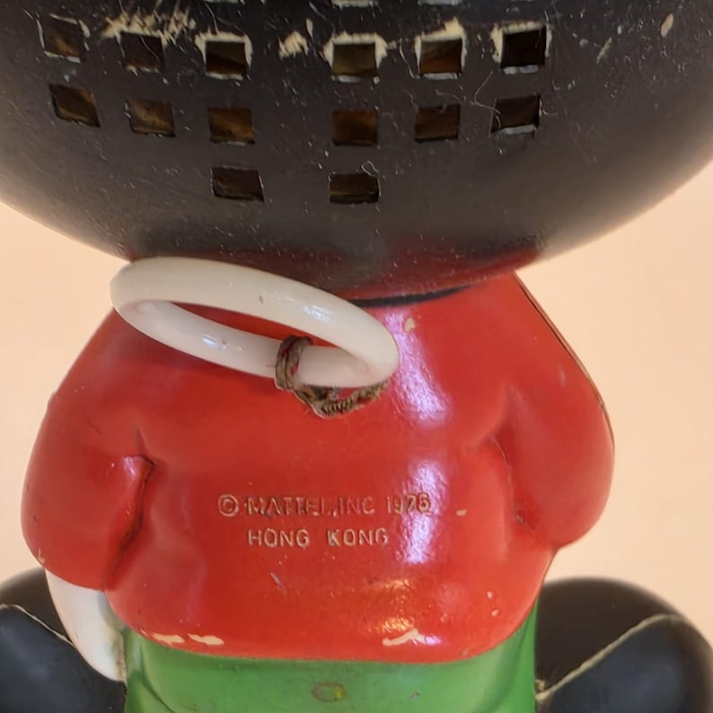 May include: Vintage toy figure with a black head and a red jacket. The figure has a white ring around its neck and green pants. The text "&copy; MATTEL, INC. 1976 HONG KONG" is printed on the red jacket.