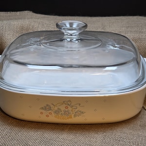 May include: A white rectangular ceramic casserole dish with integrated side handles and a clear glass lid. The front of the dish features a small, centered floral basket graphic in muted blue, green, and orange tones with tiny star accents.