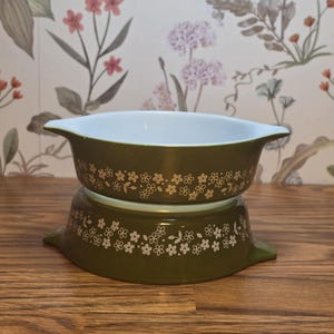May include: Two olive green Pyrex bowls with white floral patterns. The bowls have a white interior and are stacked on top of each other. The bowls are on a wooden surface with a floral wallpaper background.