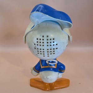 May include: A vintage Donald Duck figurine, featuring a blue sailor hat, a white head with square cutouts, and a blue and yellow sailor suit. The base is orange. The figurine is a collectible item.