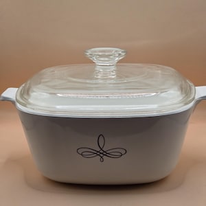 May include: A white rectangular casserole dish with integrated side handles and a clear glass lid. The front of the dish features a centered black decorative flourish design. The lid has a clear glass knob handle on top for easy removal.