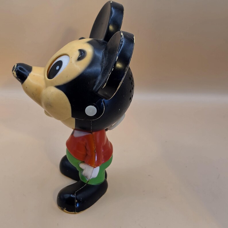 May include: Vintage Mickey Mouse toy figurine. The toy features a black head and ears, a tan face, a red shirt, green pants, and black shoes. The classic cartoon character is in a standing pose, with visible wear and tear.