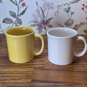 May include: Two ceramic mugs, one yellow and one white, each with a curved handle. The mugs are displayed on a wooden surface, with a floral patterned wallpaper in the background.